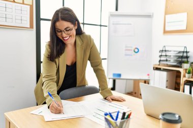 Young beautiful hispanic woman business worker writing on document standing at office