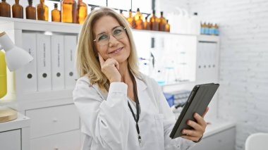 Laboratuvar önlüklü olgun kadın bilim adamı bir eczane laboratuarında elinde tablet tutarken gülümsüyor..