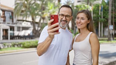 Kendine güveni tam bir baba-kız güzel bir anı paylaşıyorlar, neşeli bir şekilde cep telefonlarında serin bir selfie çekiyorlar hareketli bir şehir caddesinde duruyorlar..