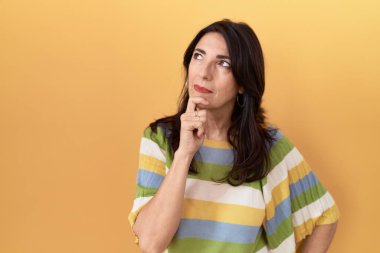 Orta yaşlı İspanyol bir kadın sarı arka planda duruyor kameraya kendinden emin bakıyor kollarını kavuşturmuş ve çenesini kaldırmış şekilde gülümsüyor. Olumlu düşünmek. 