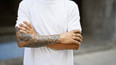 Güneşli bir caddede kolları çapraz duran kendinden emin genç Latin adam.