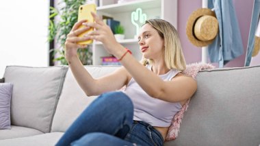 Young blonde woman taking selfie picture with smartphone sitting on the sofa at home