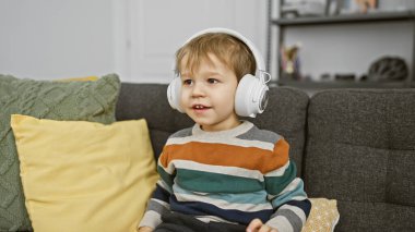 Kulaklık takan mutlu bir çocuk, rahat bir oturma odasındaki renkli yastıklarla çevrili gri bir koltukta oturur..