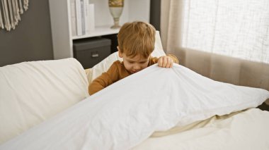 Sarışın bir çocuk, beyaz çarşaflarla güzel aydınlatılmış bir yatak odasında oynuyor..