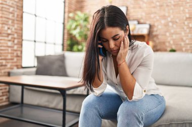 Young beautiful hispanic woman talking on smartphone with worried expression at home