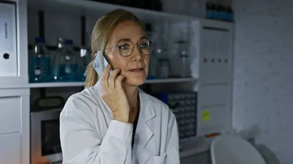 A mature caucasian woman in a lab coat talks on the phone in a laboratory setting.