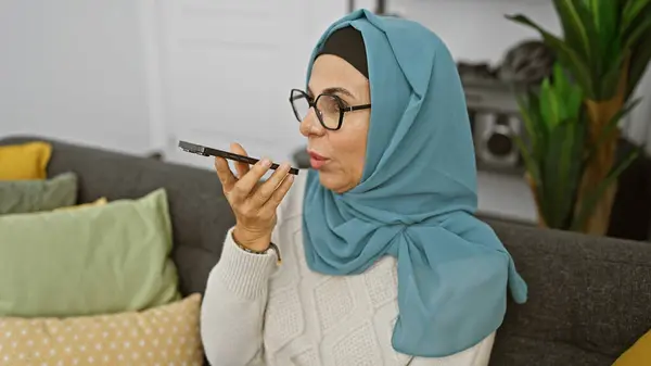 一个戴眼镜 头戴头巾的成熟女人在舒适的客厅里对着智能手机说话 — 图库照片