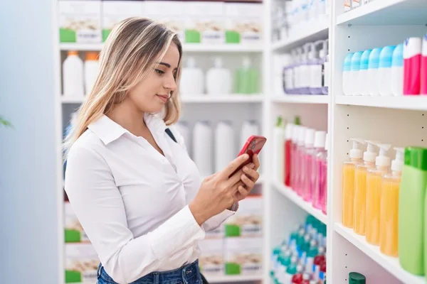 Young blonde woman customer smiling confident using smartphone at pharmacy