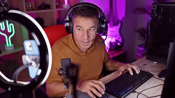 Middle-aged man in a gaming room at night conducting a videocall with headset and microphone.