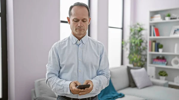 Cool hispanic middle age man in casual fashion concentrated on texting a serious message using smartphone at home, relaxed while standing in room bathed in sunlight. mature male embracing technology.