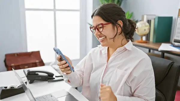 テクノロジーやパーソナルアイテムに囲まれたオフィスデスクでスマートフォンを使用してメガネでヒスパニックの女性を笑顔に — ストック写真