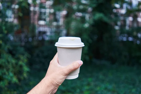 Man holding a takeaway coffee cup, with a blurred natural background suggesting an urban park.