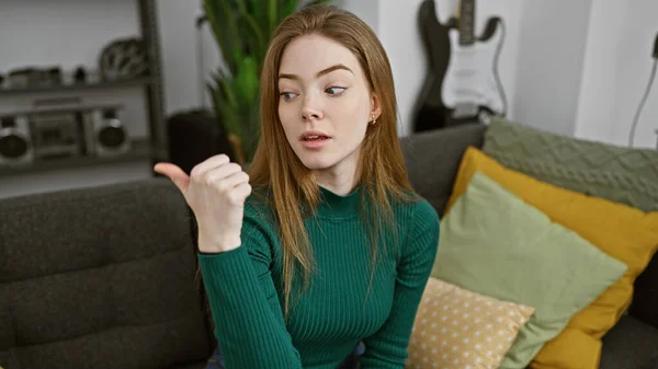 一位穿着绿色毛衣的金发女青年坐在室内 用她的大拇指摆弄着姿势 四周是一间舒适的客厅装饰着 — 图库照片