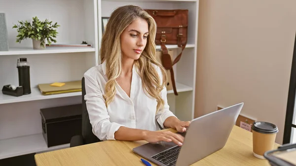 Charismatic young blonde woman boss concentrates, working online with laptop at office desk