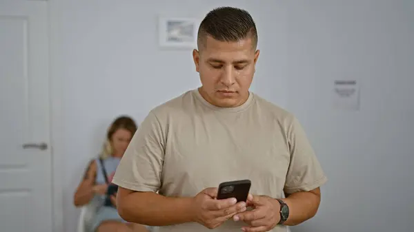 Handsome young latin man lost in his world of mobile communication, typing messages while standing in a waiting room, his face reflecting the glow of the online universe.