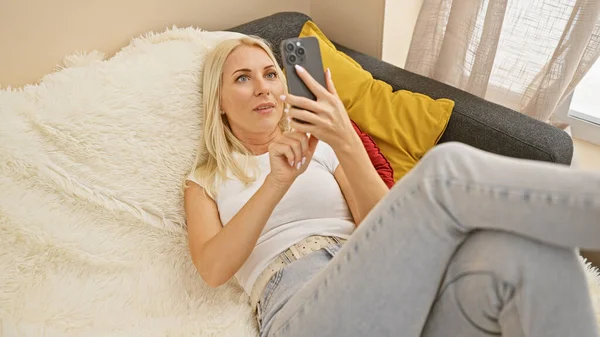 Mesajlaşmaya dalmış, kanepeye uzanmış akıllı telefonunu kullanan güzel sarışın bir kadın. İç mekan portresi, onun ciddi yüz ifadesini oturma odasında yakalıyor..