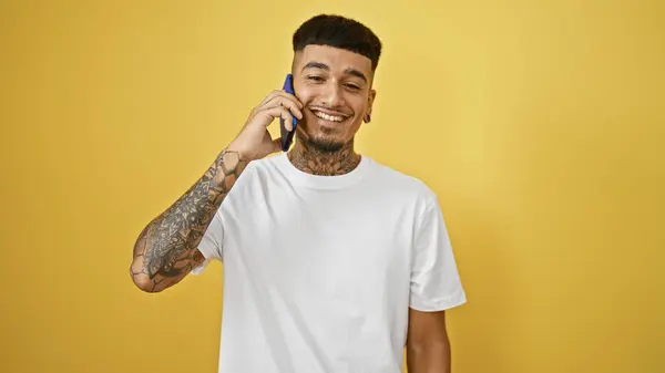 stock image Cheerful young latino man caught mid-conversation on his phone, all smiles against a vibrant yellow wall, technology sparking joy in solitude