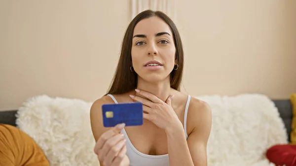 Attractive young hispanic woman lounging on living room sofa, holding credit card with serious expression, contemplating financial payment decisions indoors at home.