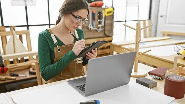 A woman wearing safety glasses and an apron taking notes on a digital tablet in a carpentry workshop with woodworking tools and a laptop on the desk.