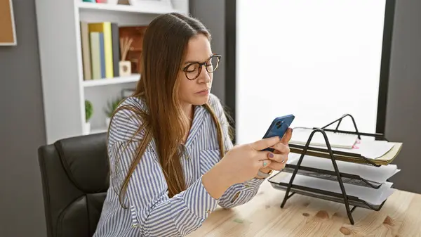 Modern ofis ortamında akıllı telefon kullanan odaklanmış bir kadın, teknoloji ve iş hayatını gözler önüne seriyor..