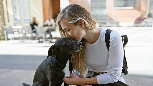 A blonde woman affectionately kisses a black dog on an urban street with blurred background.