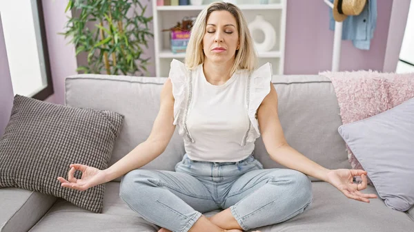 Young blonde woman doing yoga exercise sitting on sofa with serious face at home