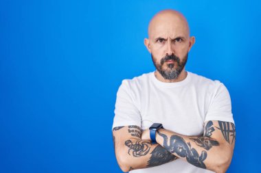 Mavi arka planda duran İspanyol dövmeli adam şüpheci ve gergin, yüzünde çapraz kollu tasvip etmeyen ifade. negatif kişi. 