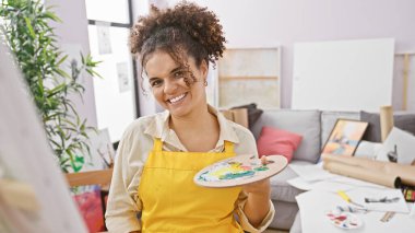 Kıvırcık saçlı, gülümseyen İspanyol kadın yaratıcı stüdyo ortamında boya paleti tutuyor..