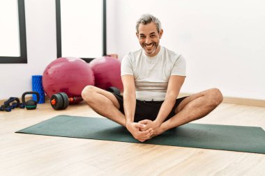 Orta yaşlı, gri saçlı, gülümseyen, spor merkezinde bacaklarını esneten bir adam.