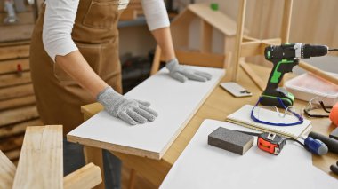 Atölyede elinde matkap ve güvenlik eldiveni olan bir kadın marangozluk yaptığını gösteriyor..