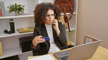 Endişeli genç bir kadın, iş yerindeki masasında konuşurken elinde bir kredi kartı tutuyor..
