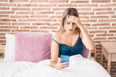 Young blonde woman using smartphone with surprise expression at bedroom