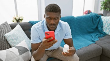 Akıllı telefon kullanan ve modern bir oturma odasında ilaç tutan Afrikalı Amerikalı bir adam..