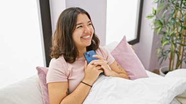 Gülümseyen genç İspanyol kadın, kapalı bir odada akıllı telefon tutuyor..