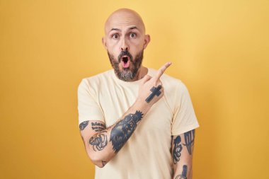 Sarı arka planda dikilen İspanyol dövmeli adam parmağını yana çevirerek şaşırdı, ağzı açık hayret içinde kaldı.. 