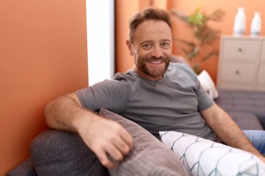 Orta yaşlı bir adam evdeki koltukta otururken kendinden emin gülümsüyor.