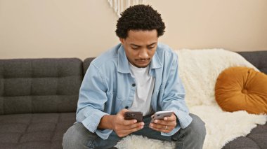 Modern bir oturma odasında otururken iki tane akıllı telefon kullanan genç bir adam..