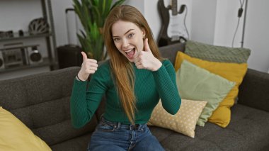 Sarı saçlı neşeli genç bir kadın evdeki pozitifliği ve mutluluğu sembolize eden rahat bir oturma odasında başparmak kaldırıyor..