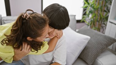 Kendine güvenen anne ve kızı, evdeki oturma odasındaki kanepede otururken içten bir gülümsemeyle neşeli bir kucaklaşmayı paylaşıyorlar. Aile sevgisinin ve mutluluğunun güzel bir ifadesi. 