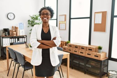 Afro-Amerikalı iş kadını gülümseyen, kendine güvenen, kollarını kavuşturup ofiste el kol hareketi yapan.