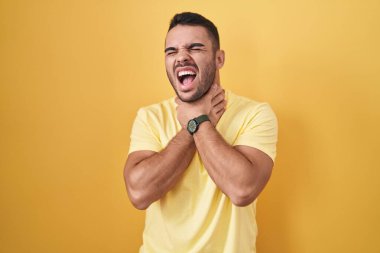 Young hispanic man standing over yellow background shouting suffocate because painful strangle. health problem. asphyxiate and suicide concept. 