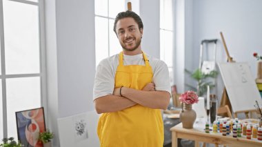 Kollarını kavuşturup sanat stüdyosuna gülümseyen genç İspanyol sanatçı.