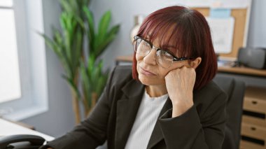 Düşünceli orta yaşlı, gözlüklü İspanyol kadın modern ofis ortamında tefekkür ediyor..