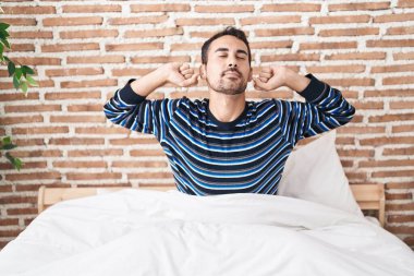 Young hispanic man waking up stretching arms at bedroom