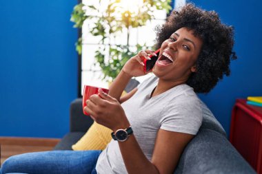 Afro-Amerikalı kadın akıllı telefondan konuşuyor evde kahve içiyor.