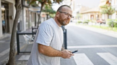 Beyaz adam sokakta ciddi bir ifadeyle akıllı telefon kullanıyor.