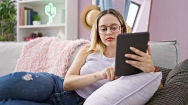Young blonde woman using touchpad sitting on sofa at home
