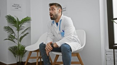 Aşırı çalışan İspanyol asıllı genç bir adam, üniformalı stresli bir doktor kliniğin bekleme odasında ciddi bir şekilde oturuyor, sağlık hizmetleri stresinin bedelini gösteriyor.