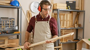 A young hispanic man wearing safety equipment examines lumber in a well-organized carpentry workshop. clipart