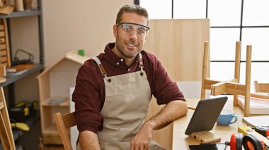 Güvenlik gözlüğü takan ve marangozluk atölyesinde aletleri ve ahşap mobilyaları olan gülümseyen sakallı bir adam..